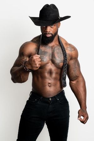 Shirtless muscular fitness model with beard and tattoos wearing a black cowboy hat and dark jeans, gripping suspenders in a studio portrait.