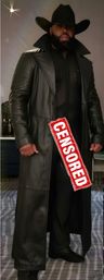 Person wearing a wide-brim black cowboy hat, full beard and long black leather trench coat over a sheer black outfit, red "CENSORED" stamp across the front, standing indoors on a patterned carpet.