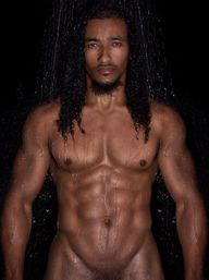 Studio portrait of a shirtless male fitness model with long wet curls and defined abs, water streaming down a glistening torso against a black background