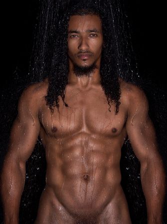 Studio portrait of a shirtless male fitness model with long wet curls and defined abs, water streaming down a glistening torso against a black background