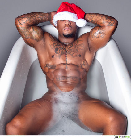 Tattooed muscular man reclining in a white bathtub with a Santa hat pulled over his eyes, relaxing in a bubbly holiday studio soak