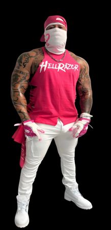 Muscular tattooed person in bright pink sleeveless top, white face covering and cap, pink gloves, white pants and boots standing against a black background.