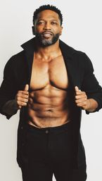 Studio portrait of a fit male model holding open a black shirt to reveal sculpted six-pack abs and a toned chest, wearing black pants against a clean white background.