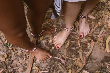 Two barefoot people wearing red-and-white beaded anklets standing on a leaf-strewn forest floor; one foot has red toenail polish and an ankle tattoo