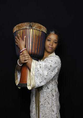 Studio portrait of a woman cradling a large West African djembe drum against a black background, wearing a gold-and-white patterned robe and red-and-white beaded bracelets.