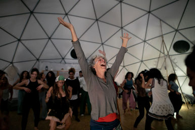 Joyful gray-haired person with arms raised, cheering in the center of a lively dance circle inside a geodesic dome, surrounded by people dancing and laughing.
