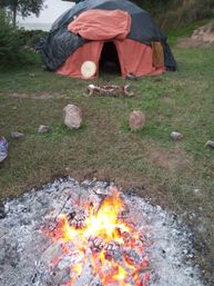 Glowing campfire in foreground with a small handmade yurt draped in orange and black fabric, drum and arranged stones on a grassy outdoor clearing