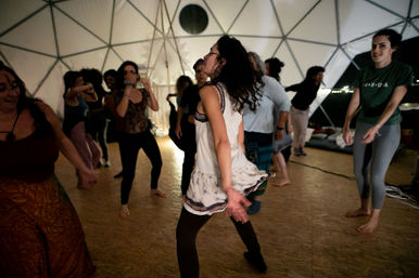 People dancing barefoot inside a lit geodesic dome, a person in a white dress twirling in the foreground during a lively community dance event on a wooden floor.