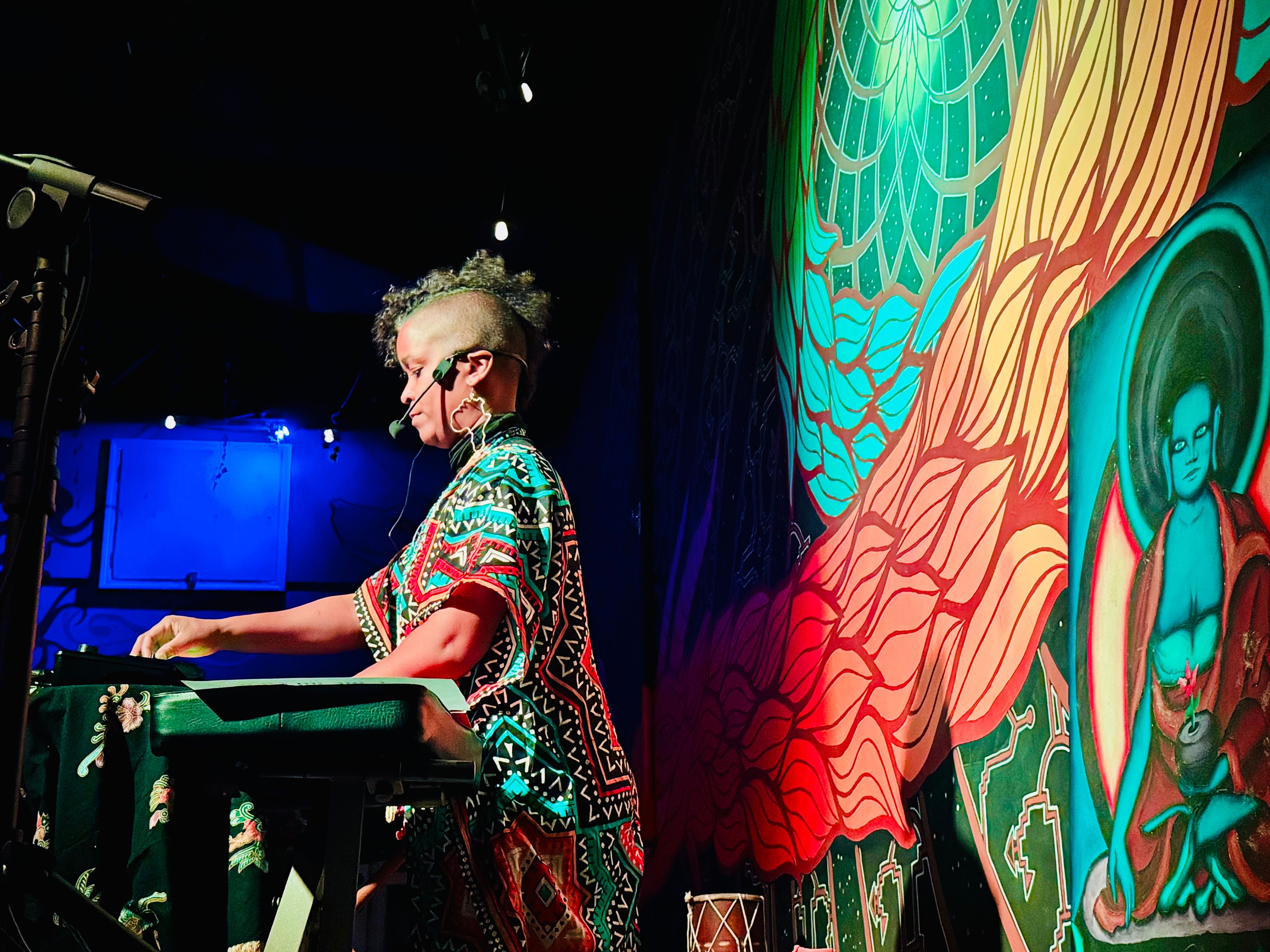 Performer with headset playing a synthesizer at an indoor live-music venue, colorful mural backdrop with a meditative figure, bold red and blue stage lighting, funky patterned outfit and shaved-sided mohawk.