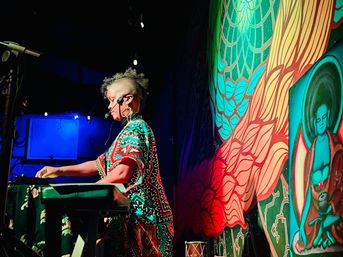 Performer with headset playing a synthesizer at an indoor live-music venue, colorful mural backdrop with a meditative figure, bold red and blue stage lighting, funky patterned outfit and shaved-sided mohawk.