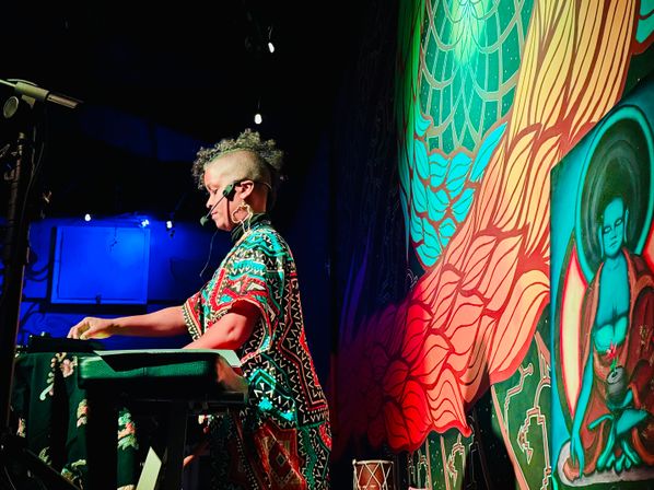 Performer with headset playing a synthesizer at an indoor live-music venue, colorful mural backdrop with a meditative figure, bold red and blue stage lighting, funky patterned outfit and shaved-sided mohawk.