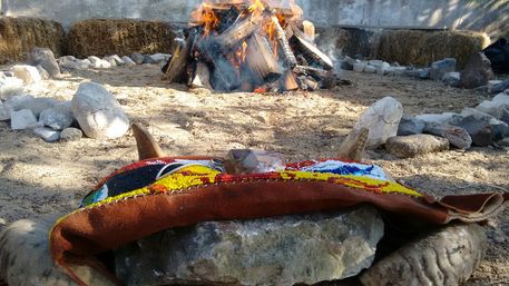 Colorful beaded leather pouch with a crystal and small antler resting on a stone in front of a wood bonfire inside an outdoor fire circle surrounded by rocks and hay bales.