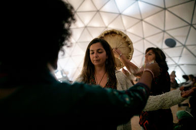 Meditative woman with eyes closed in a group drum circle inside a geodesic dome, participants with outstretched arms and a hand‑held frame drum overhead.
