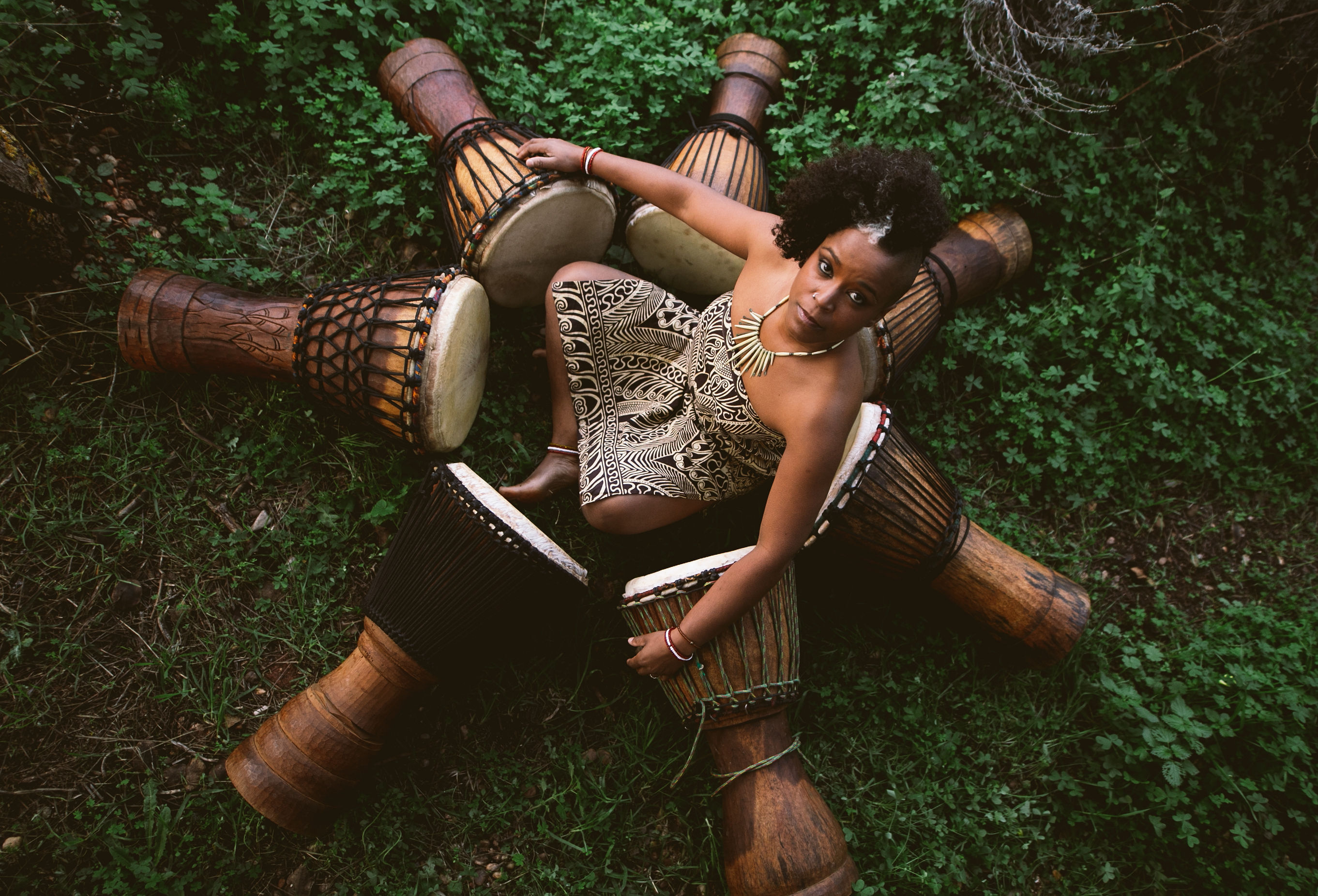 Overhead view of a woman seated outdoors on green grass, surrounded by a circle of wooden djembe drums, wearing a patterned dress and statement necklace, looking up at the camera.