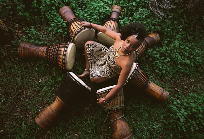 Overhead view of a woman seated outdoors on green grass, surrounded by a circle of wooden djembe drums, wearing a patterned dress and statement necklace, looking up at the camera.