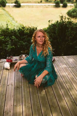 Woman in a teal lace dress with curly blonde hair sitting cross-legged on a wooden deck by green shrubs, with a small tray of candles and crystals and a large seashell nearby and a grassy countryside field in the background.