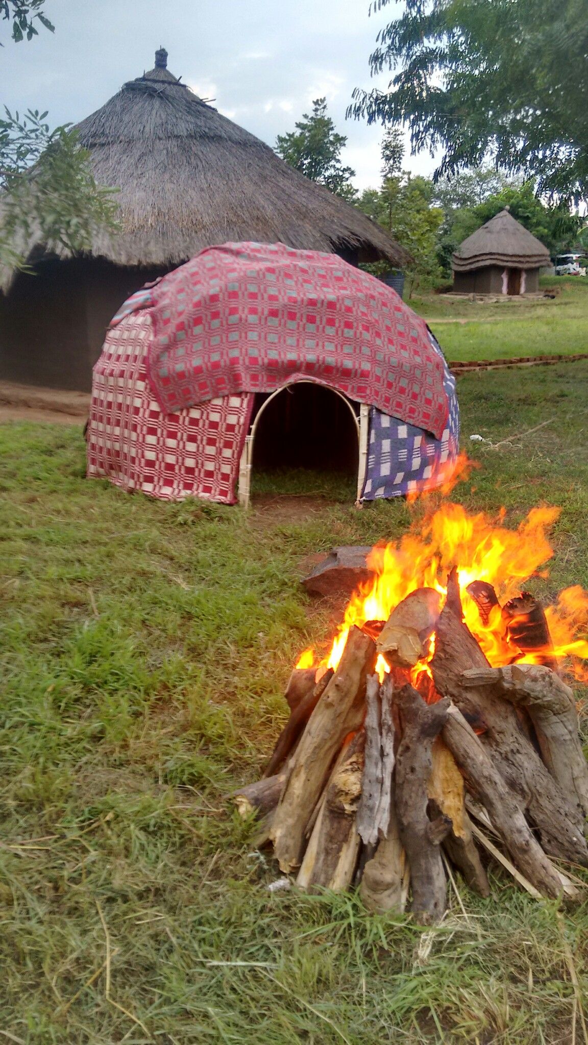 Blazing bonfire of stacked logs on a grassy clearing with a colorful fabric-covered dome and traditional round thatched huts in a rural village under a cloudy sky.