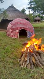 Blazing bonfire of stacked logs on a grassy clearing with a colorful fabric-covered dome and traditional round thatched huts in a rural village under a cloudy sky.