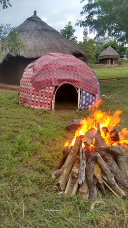 Blazing bonfire of stacked logs on a grassy clearing with a colorful fabric-covered dome and traditional round thatched huts in a rural village under a cloudy sky.
