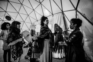 Black-and-white photo of a small group inside a geodesic dome participating in a sound-healing circle — one woman plays a hand drum, another plays a flute, a central woman stands with open arms.
