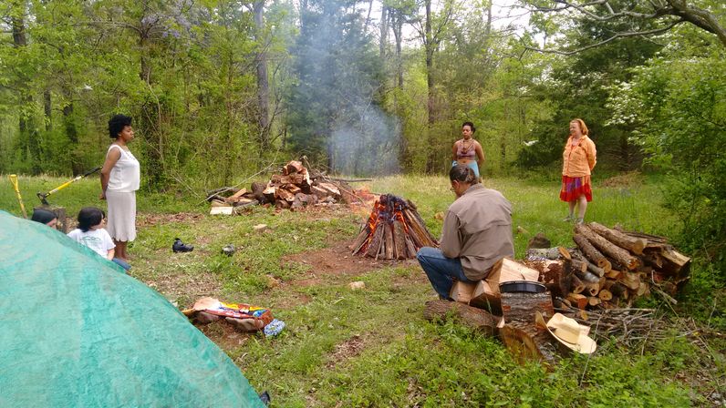 Group camping in a forest clearing around a smoking bonfire, with stacked firewood, logs and a green tent nearby — outdoor woodland gathering in spring.