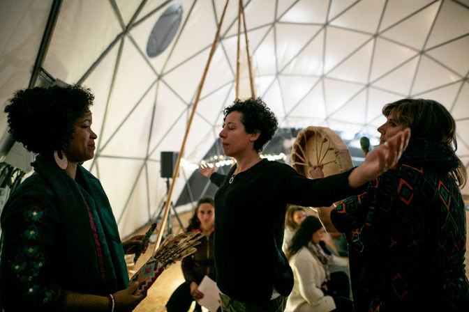 Three participants in a geodesic dome engage in a community wellness circle, one person standing with arms outstretched while others hold a hand drum and feather fan during a ceremonial drumming/smudging moment.