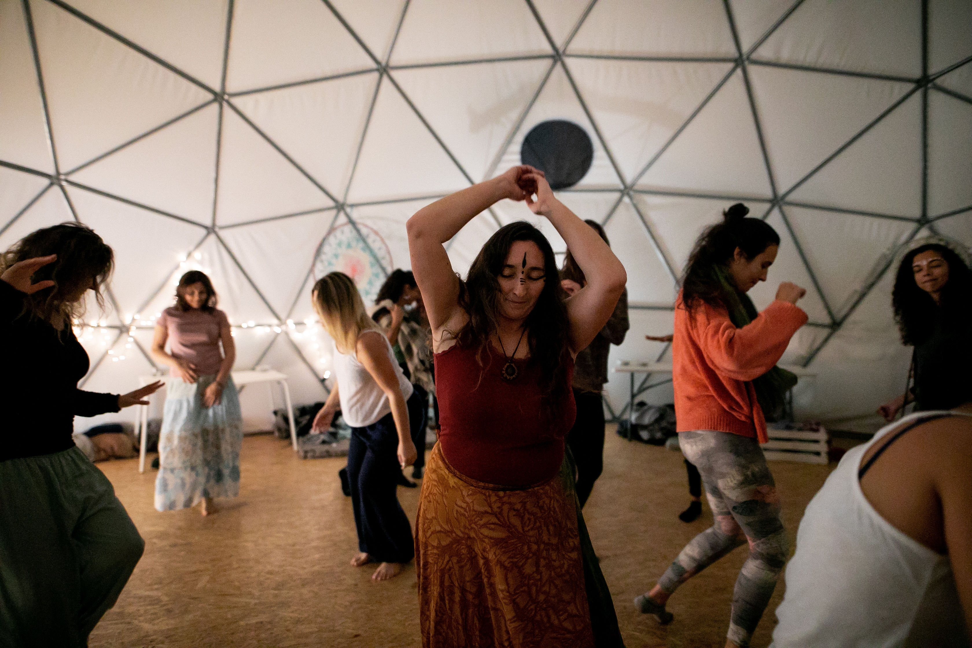 Barefoot community dancing in a cozy geodesic dome, woman in a red top leading a joyful movement circle.