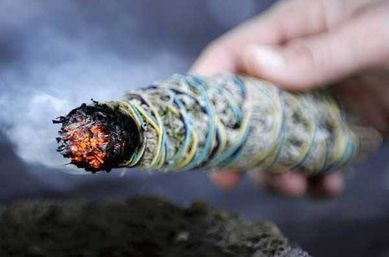 Hand holding a lit sage smudge stick wrapped in colorful thread, glowing ember and curling smoke for a cleansing ritual