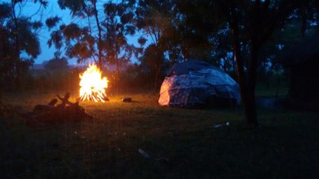 Dusk campsite in a tree-lined clearing with a glowing bonfire and a tarp-covered dome tent on grassy ground