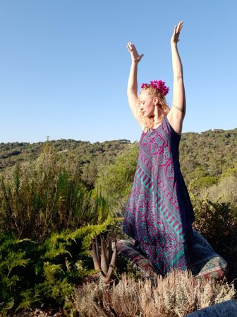Woman in a purple patterned boho dress and pink flower crown standing on cushions with arms raised on a sunny wooded hillside under a clear blue sky