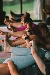 Row of people in a sunlit yoga studio lying on mats with legs extended and hands holding feet during a group stretching pose.
