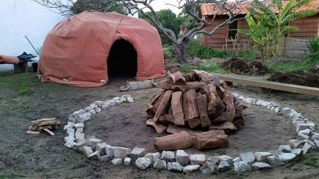 Outdoor backyard earthen oven draped in orange canvas with dark entrance, stacked firewood inside a circular stone fire pit, wooden shed and tropical plants in the background.