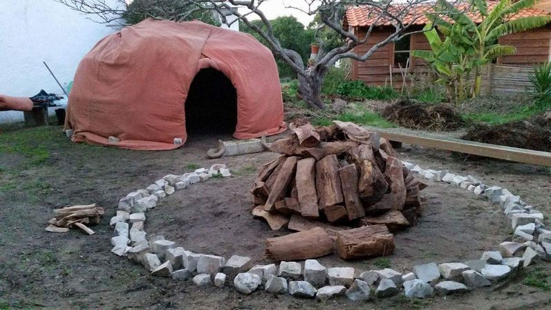 Outdoor backyard earthen oven draped in orange canvas with dark entrance, stacked firewood inside a circular stone fire pit, wooden shed and tropical plants in the background.