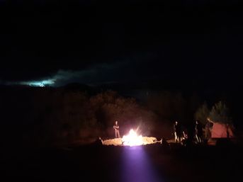 Nighttime outdoor camping scene with people gathered around a glowing bonfire, a nearby tent, brush-covered hills and a cloudy moonlit sky.