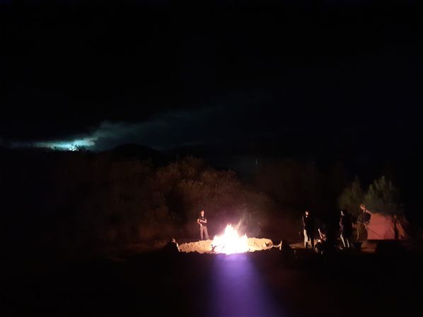 Nighttime outdoor camping scene with people gathered around a glowing bonfire, a nearby tent, brush-covered hills and a cloudy moonlit sky.