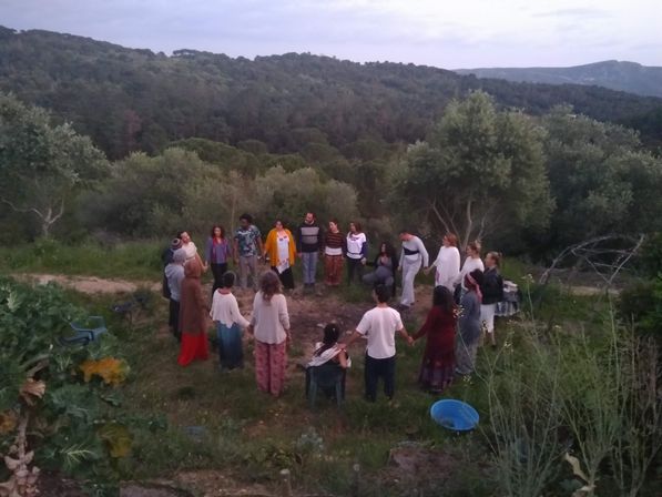 Circle of people holding hands during an outdoor gathering on a grassy hillside, surrounded by dense woodland and rolling hills.