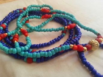 Close-up of coiled colorful beaded necklaces with turquoise and deep blue seed beads, red accent beads and small gold spacer beads on a light fabric background