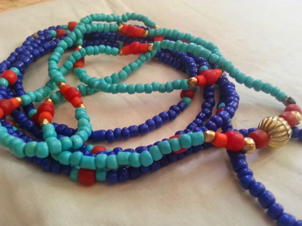 Close-up of coiled colorful beaded necklaces with turquoise and deep blue seed beads, red accent beads and small gold spacer beads on a light fabric background