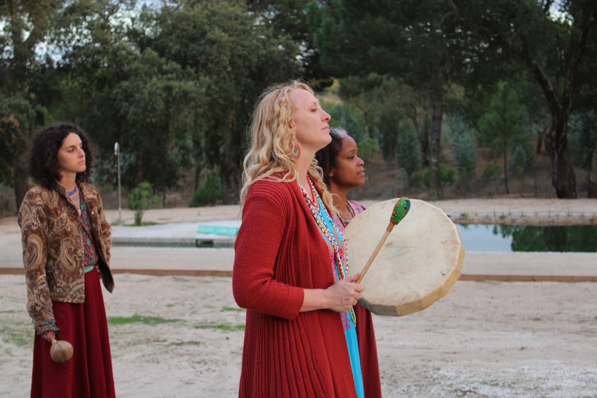 Three women in colorful bohemian outfits at a wooded lakeside — one in a red cardigan plays a round frame drum with a padded mallet while the others stand nearby holding shakers.