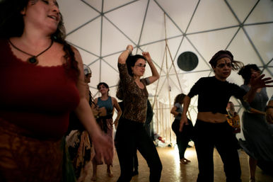 People dancing energetically inside a geodesic dome with white triangular panels, warm floor lighting, bohemian outfits and painted faces — lively indoor festival vibe