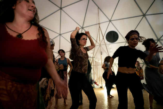 People dancing energetically inside a geodesic dome with white triangular panels, warm floor lighting, bohemian outfits and painted faces — lively indoor festival vibe