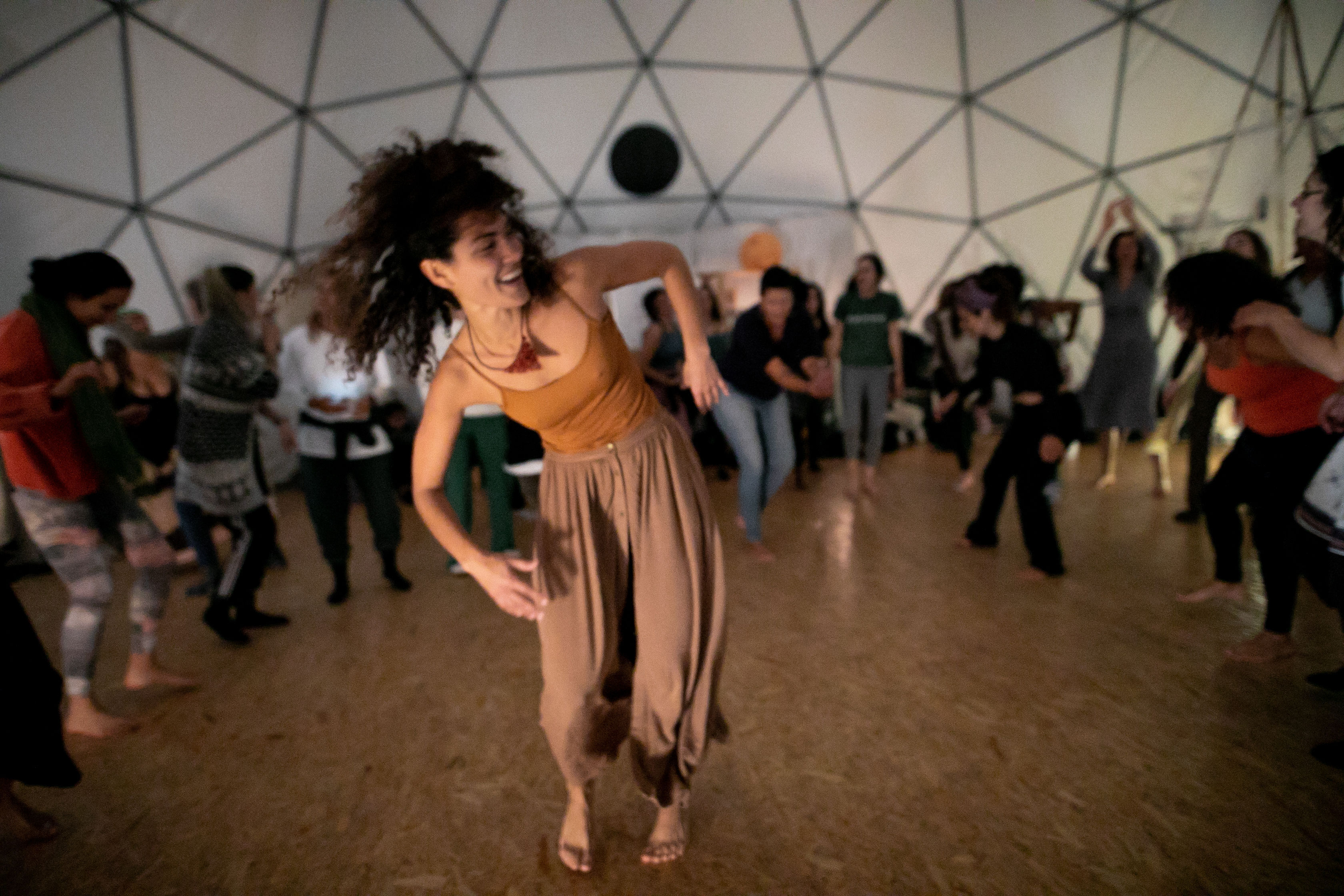 Barefoot woman dancing joyfully at the center of a lively community dance workshop inside a geodesic dome, surrounded by participants moving in a circle.