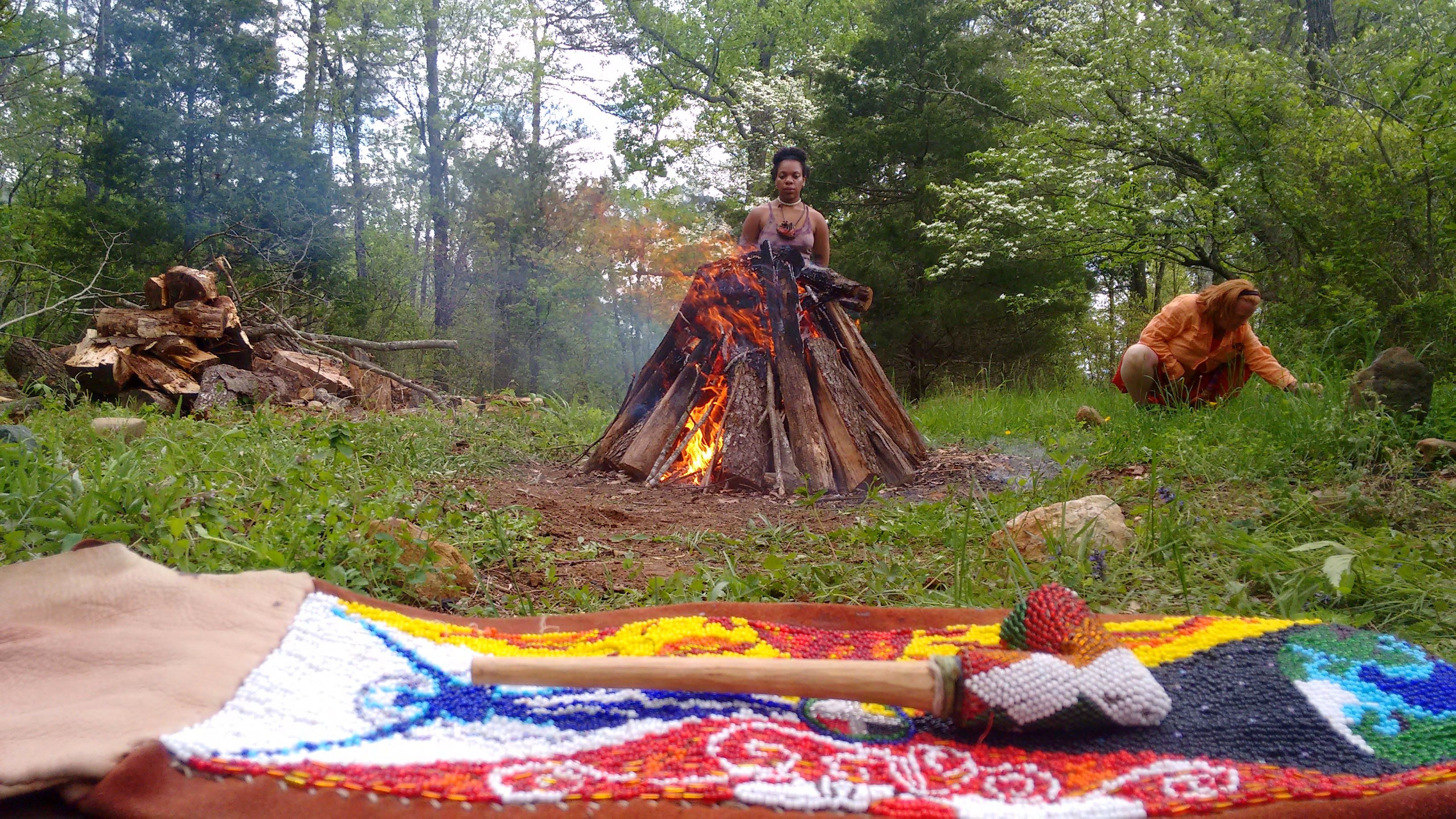 Handmade beaded rattle on a colorful textile in the foreground, teepee-style bonfire burning in a green woodland clearing with two people nearby and a pile of firewood — outdoor forest bonfire scene.