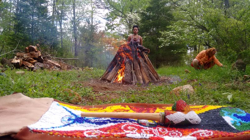 Handmade beaded rattle on a colorful textile in the foreground, teepee-style bonfire burning in a green woodland clearing with two people nearby and a pile of firewood — outdoor forest bonfire scene.