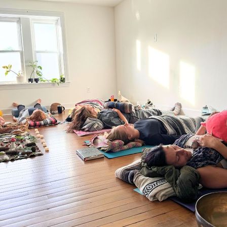 Group of people resting on mats for a guided meditation/sound-bath in a bright sunlit studio with hardwood floors, cozy blankets, pillows and window plants.