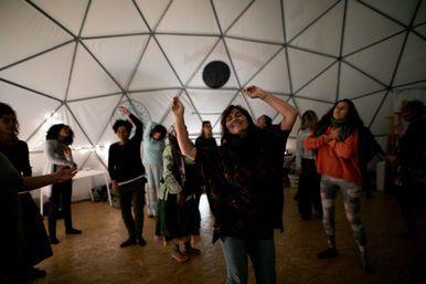 Community dance workshop inside a geodesic dome: a smiling woman in the foreground raises her arms while others move around under soft string lights and triangular dome panels.
