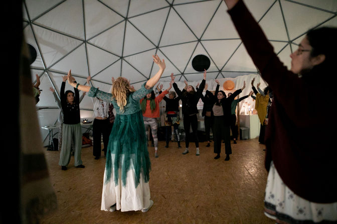 Instructor in a flowing green dress leads a standing group with arms raised inside a white geodesic dome during a community movement/wellness workshop.