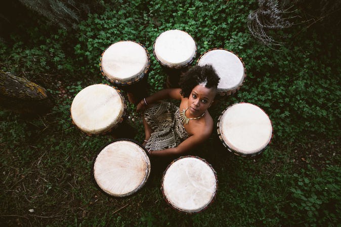 Aerial view of a woman seated in green grass surrounded by a circle of seven djembes (African hand drums), wearing a patterned wrap and statement necklace.