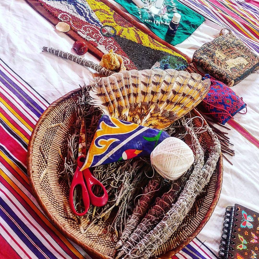 Woven basket holding smudge bundles, a striped feather fan, white twine, red scissors and colorful fabric on a striped boho blanket.