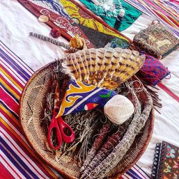 Woven basket holding smudge bundles, a striped feather fan, white twine, red scissors and colorful fabric on a striped boho blanket.
